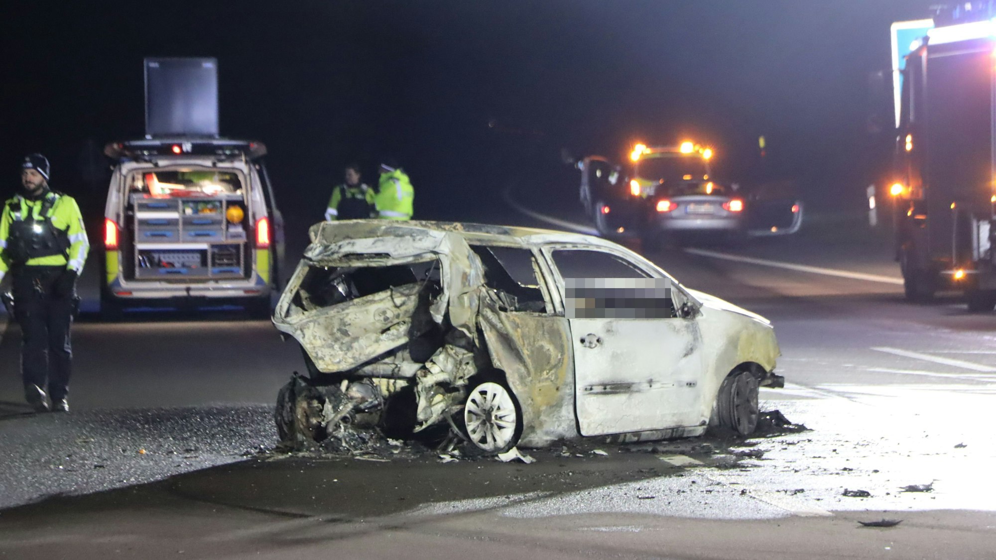 Ein Kleinwagen steht ausgebrannt und mit stark beschädigtem Heck quer auf der Autobahn, Einsatzkräfte sind vor Ort.