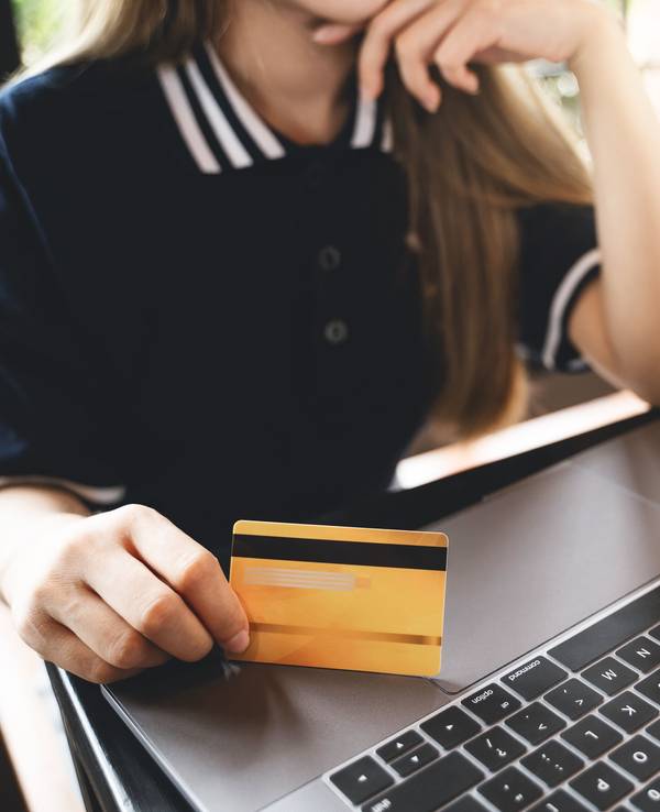 Symbolfoto: Eine Frau sitzt vor einem Laptop und hält eine Kreditkarte für das Online-Shopping bereit.
