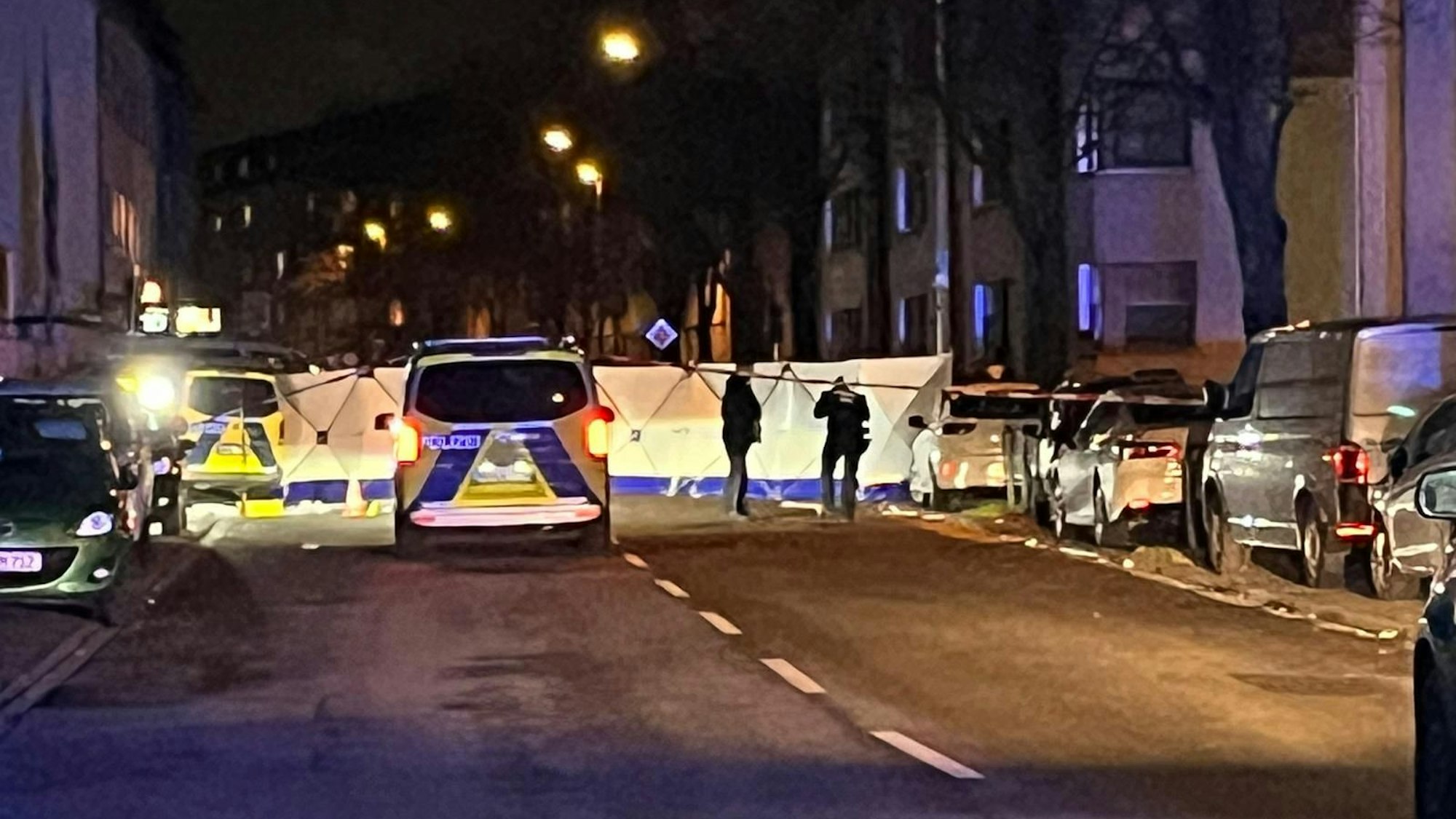 Die Polizei hat die Straße abgesperrt und einen Sichtschutz aufgebaut. Davor stehen zwei Beamte sowie ein Einsatzfahrzeug.