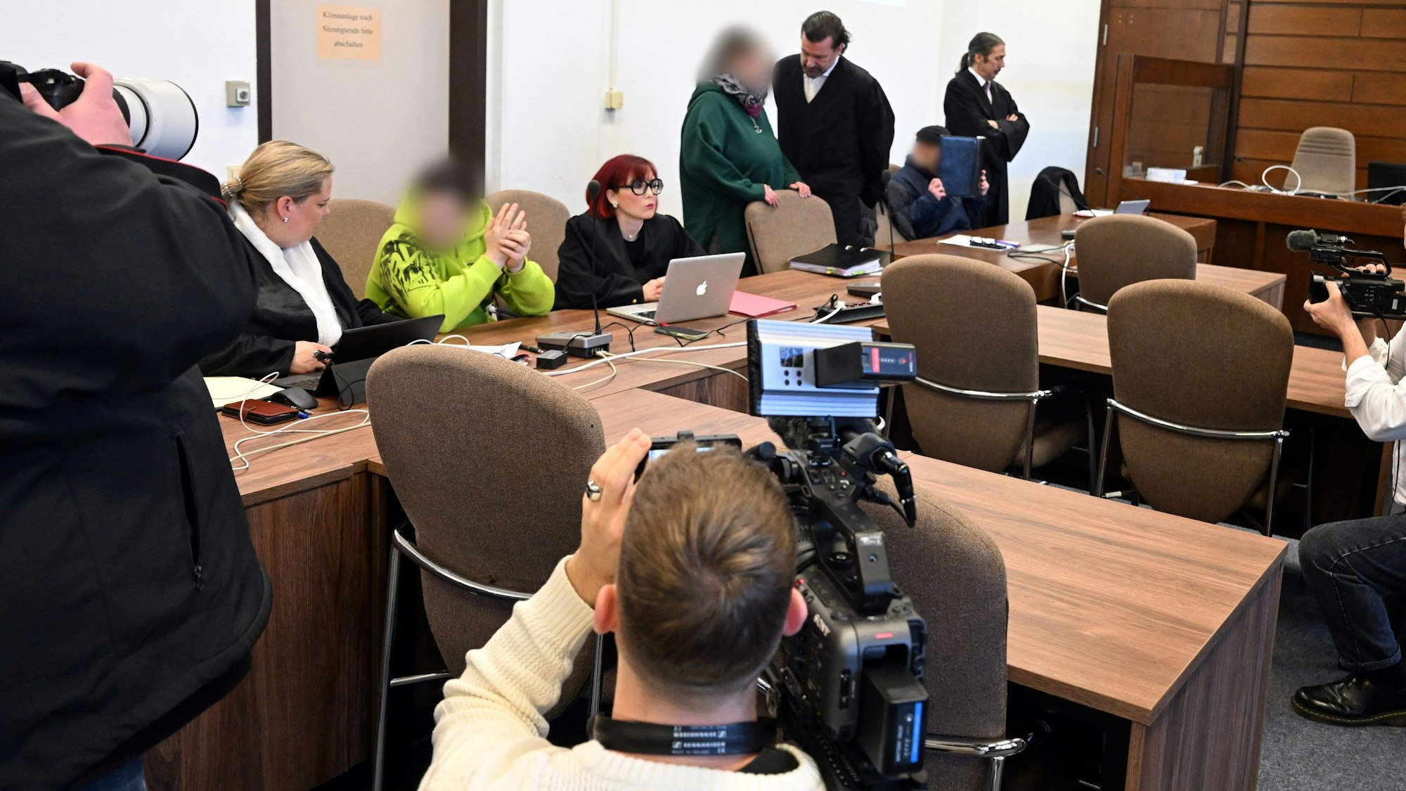 In einem Gerichtssaal warten Angeklagte und ihre Anwälte auf den Prozessbeginn, zwei Fotografen und ein Kamera-Mann halten die Szene fest.