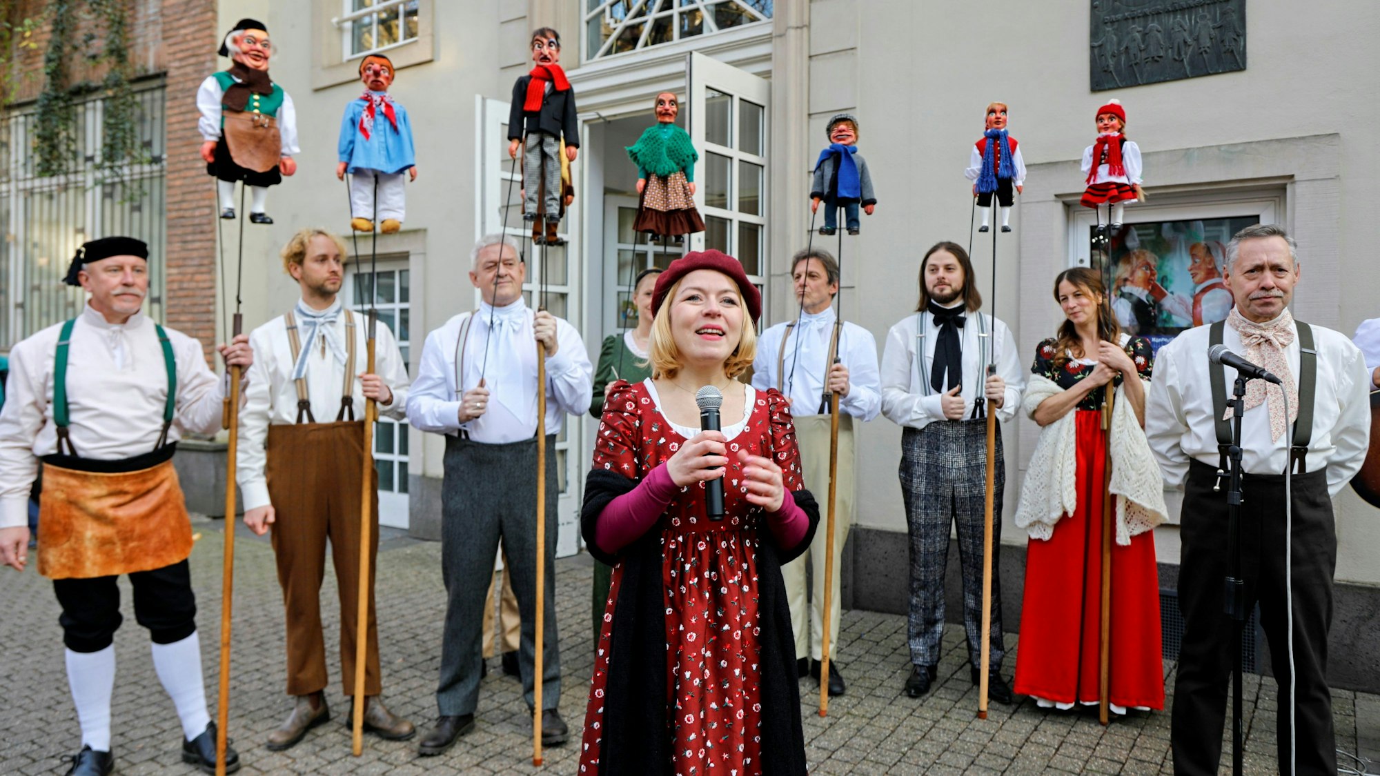 Puppenspielerinnen und -spieler stehen in einem Halbkreis. Eine Frau spricht davor in ein Mikrofon.