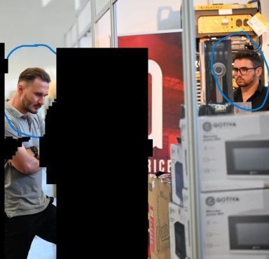 Zwei Männer stehen an einem Messestand, ihre Köpfe sind auf dem Fahndungsfoto blau eingekreist.