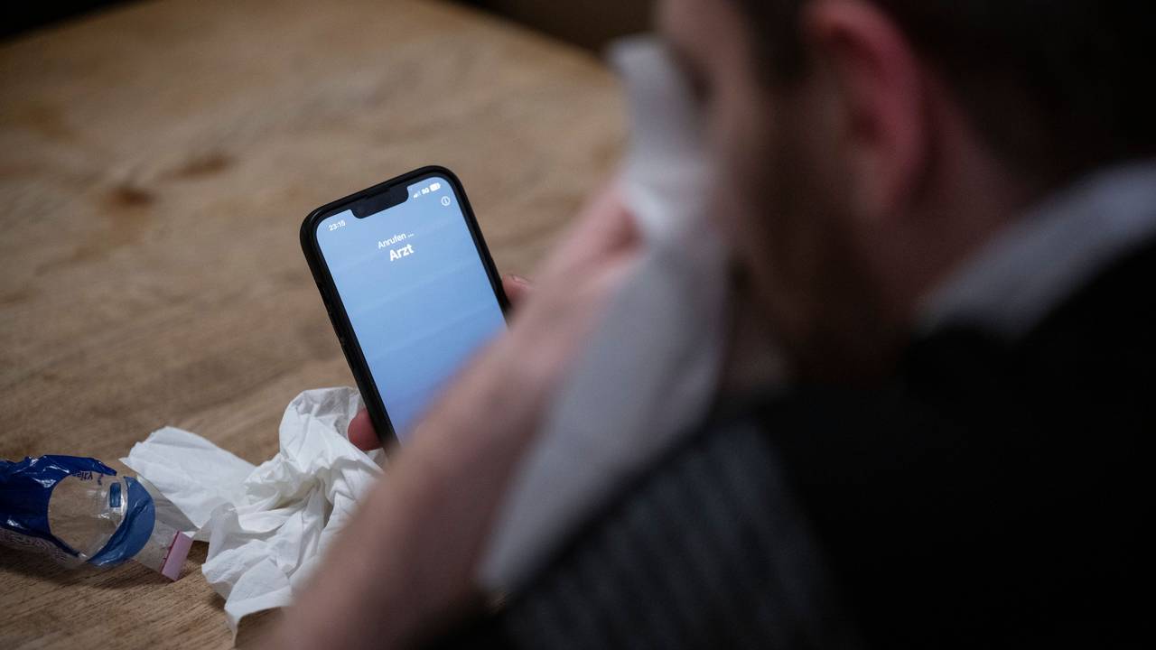 Ein Mann hält ein Taschentuch in einer Hand und ein Telefon mit dem Schriftzug „Arzt“ in der Anderen.