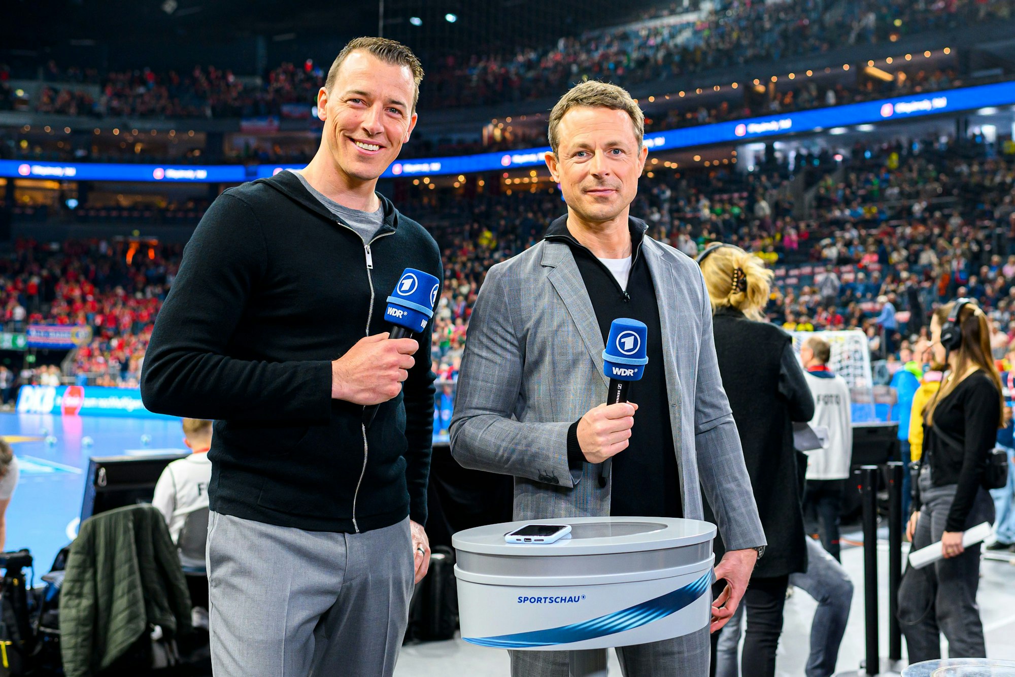 Dominik Klein und Alexander Bommes stehen am Moderatoren-Tisch und lächeln in die Kamera. Beide halten ein ARD-Mikro in der Hand.