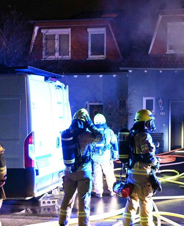 Einsatzkräfte und -fahrzeuge stehen vor einem Einfamilienhaus, aus dem Rauch dringt. Löschschläuche liegen auf dem Boden.