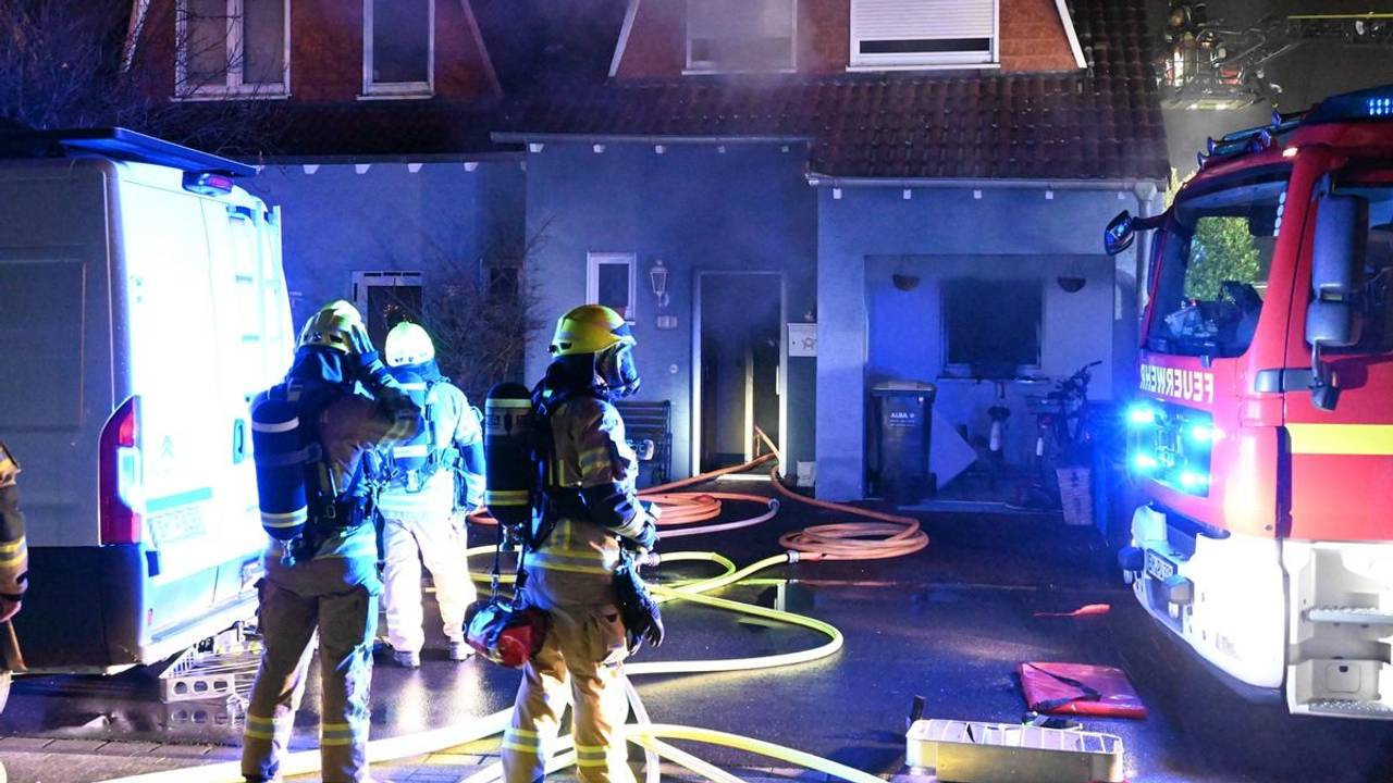 Einsatzkräfte und -fahrzeuge stehen vor einem Einfamilienhaus, aus dem Rauch dringt. Löschschläuche liegen auf dem Boden.