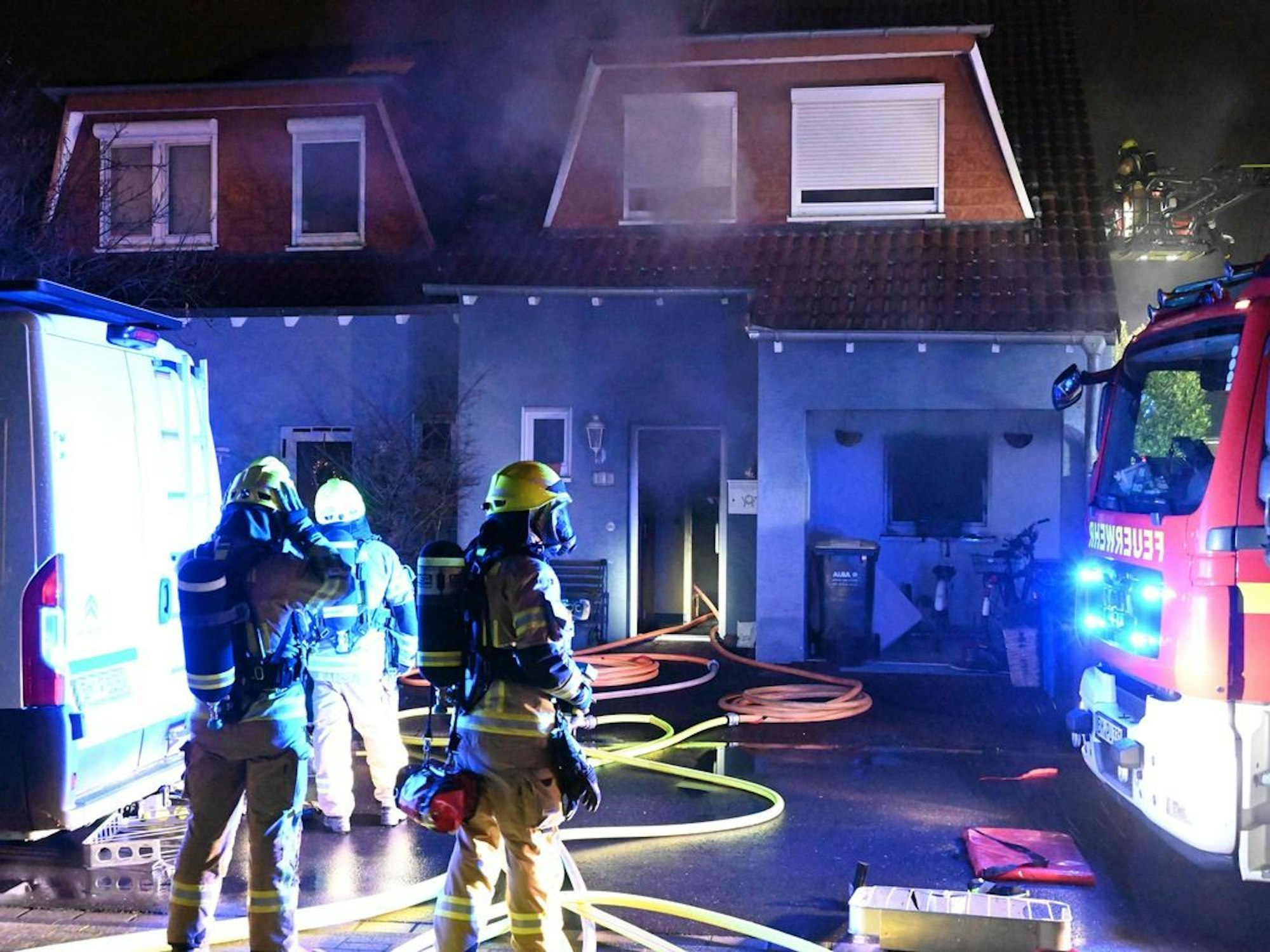 Einsatzkräfte und -fahrzeuge stehen vor einem Einfamilienhaus, aus dem Rauch dringt. Löschschläuche liegen auf dem Boden.