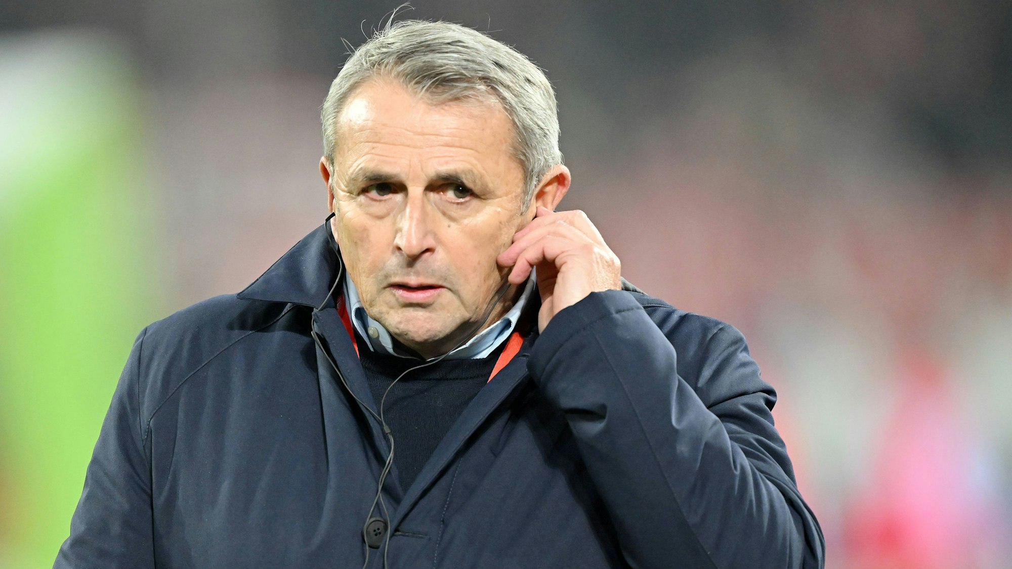 Fortuna Düsseldorfs Sport-Vorstand Klaus Allofs steht vor dem Spiel gegen den FC Schalke 04 auf dem Platz und greift sich ans Ohr.