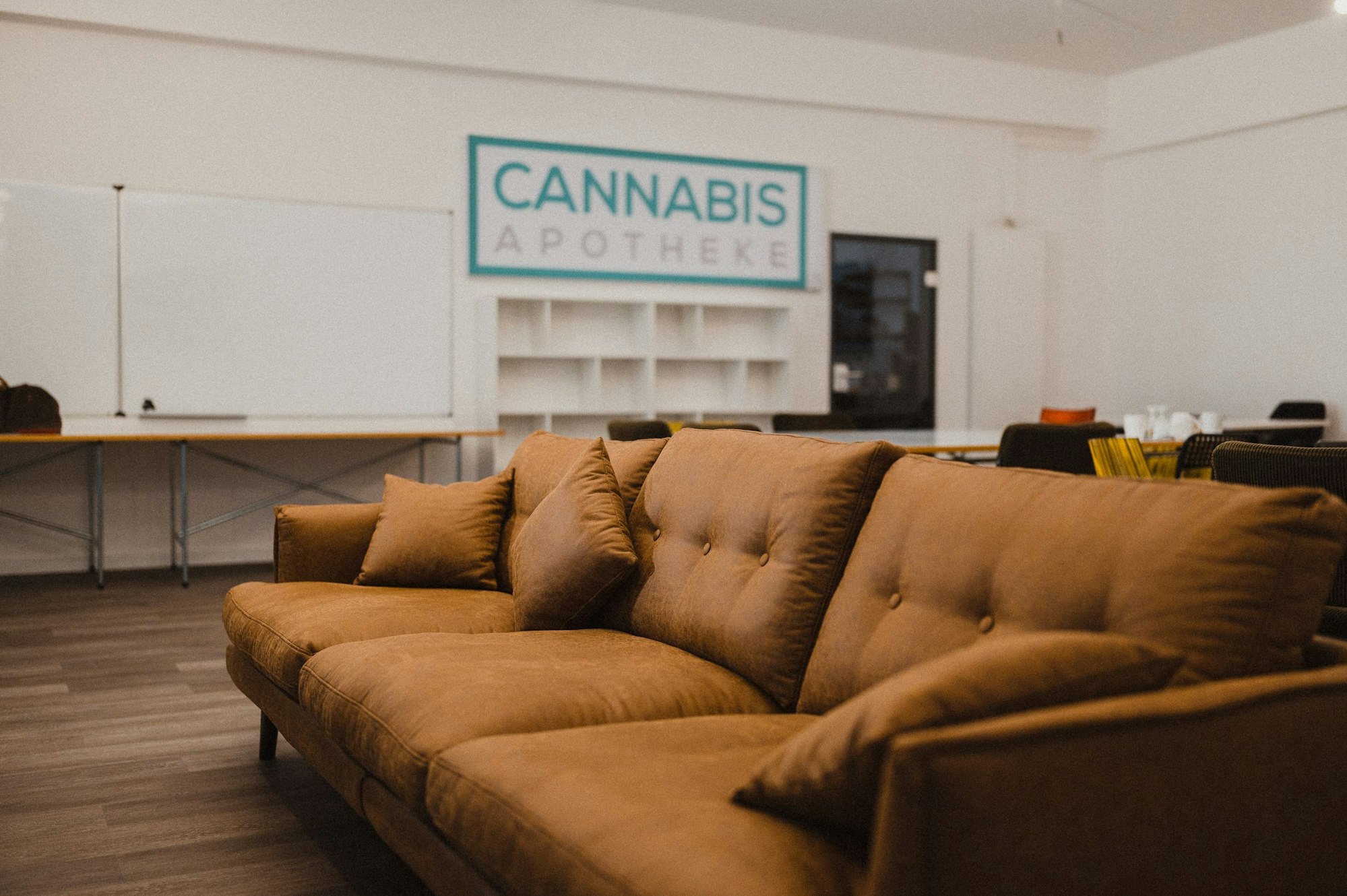 Ein Sofa in einem Seminarraum mit Tischen und Regalen im Hintergrund. Auf der Wand steht der Schriftzug “Cannabis Apotheke”.
