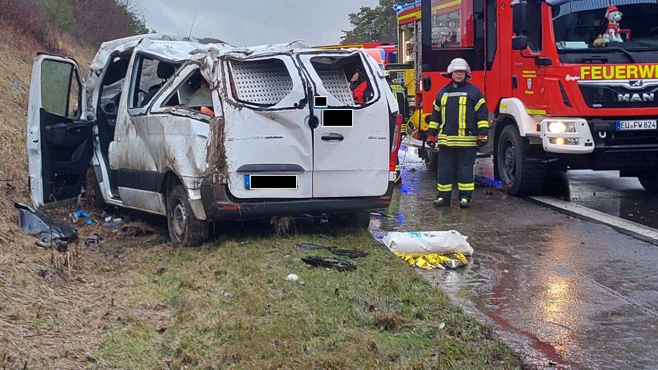Ein schwer beschädigter Kleintransporter steht neben der A1-Fahrbahn, das Dach ist stark eingedellt. Daneben steht ein Fahrzeug der Feuerwehr.