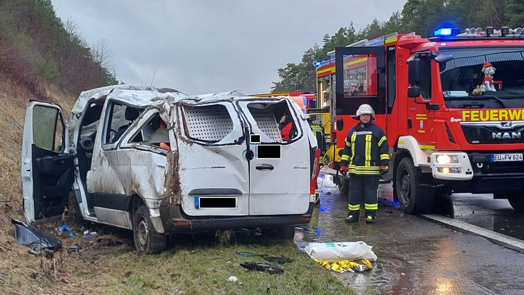 Ein schwer beschädigter Kleintransporter steht neben der A1-Fahrbahn, das Dach ist stark eingedellt. Daneben steht ein Fahrzeug der Feuerwehr.