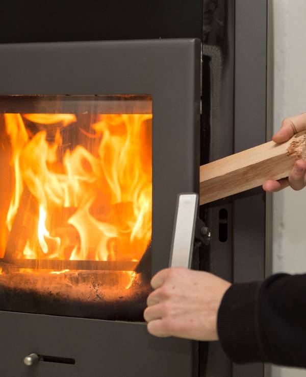 Ein Mann legt in seiner Wohnung ein Stück Holz in einen Kaminofen.