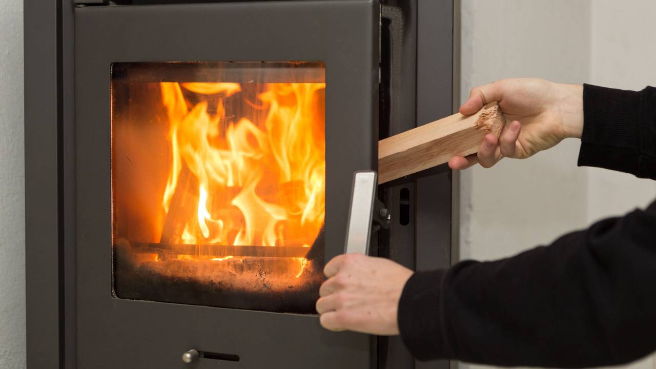 Ein Mann legt in seiner Wohnung ein Stück Holz in einen Kaminofen.