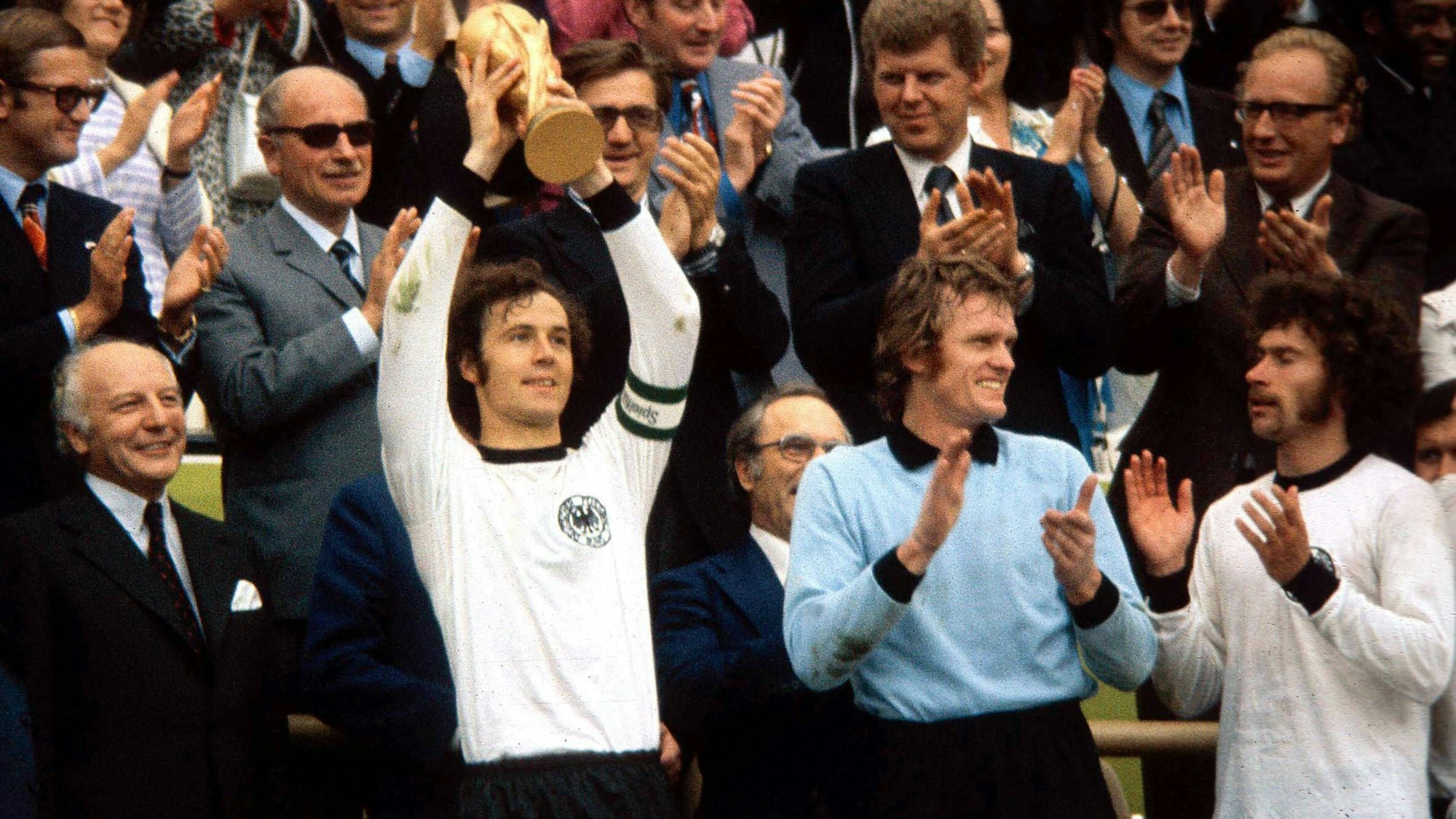 Franz Beckenbauer reckt den WM-Pokal in die Luft. Sepp Maier und Paul Breitner applaudieren.