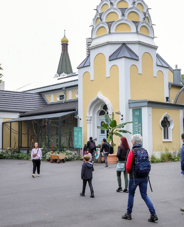 Menschen gehen durch den Kölner Zoo, im Hintergrund ist das Reichert-Haus zu sehen.