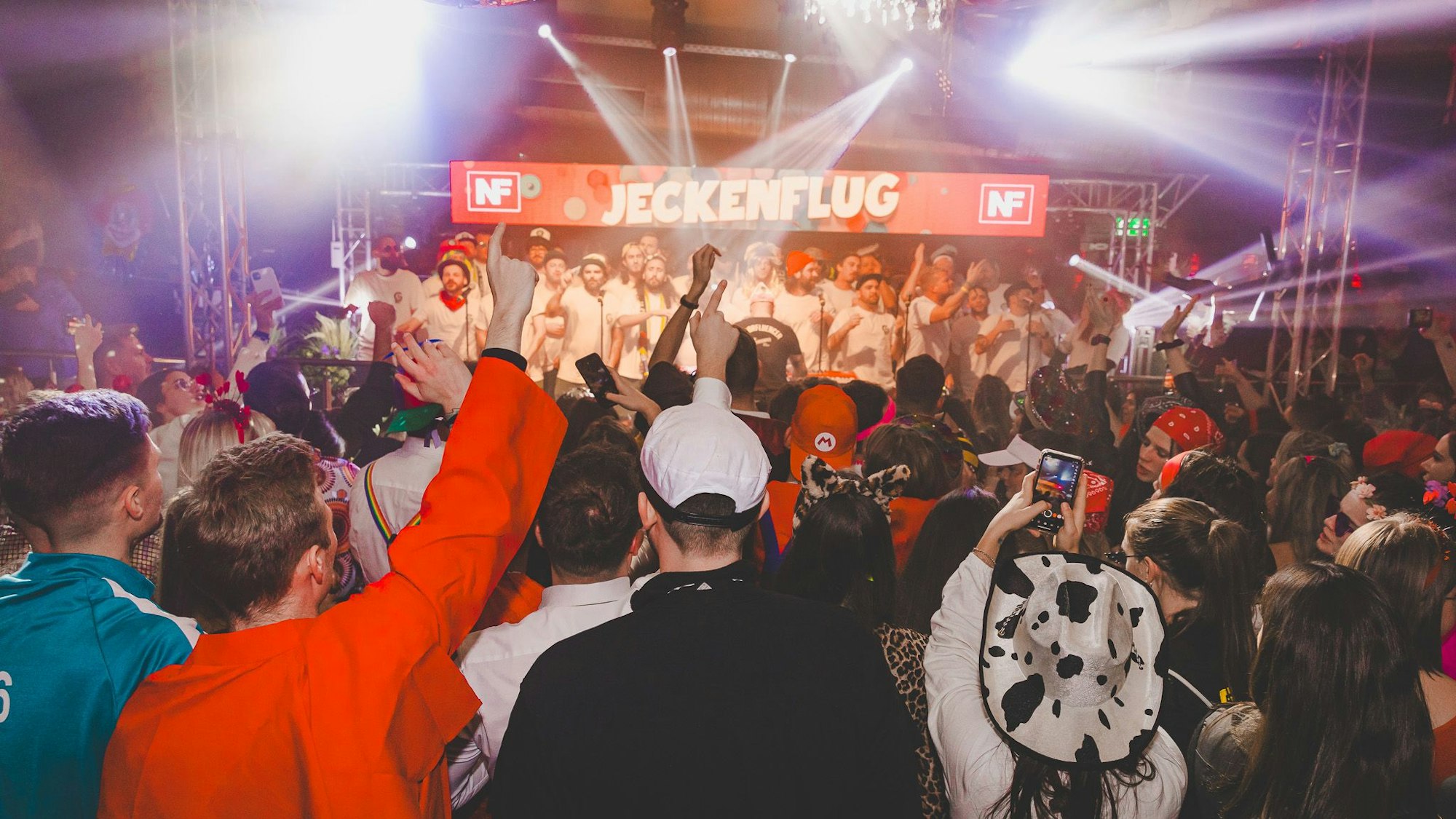 Viele kostümierte Personen stehen in einem Club vor einer Bühne, auf der eine Männergruppe singt.