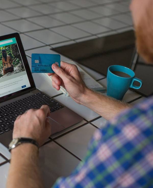 Auf dem Foto sieht man einen Mann vor einem Laptop sitzen, um ihn herum steht eine Kaffeetasse, ein Reisepass, Geld und eine Kamera.