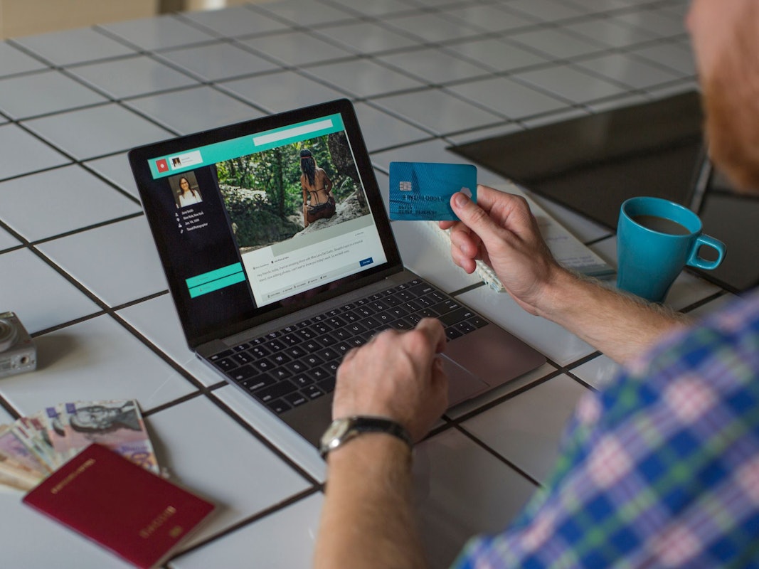 Auf dem Foto sieht man einen Mann vor einem Laptop sitzen, um ihn herum steht eine Kaffeetasse, ein Reisepass, Geld und eine Kamera.