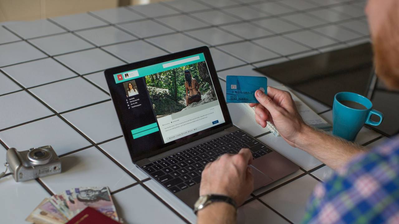 Auf dem Foto sieht man einen Mann vor einem Laptop sitzen, um ihn herum steht eine Kaffeetasse, ein Reisepass, Geld und eine Kamera.