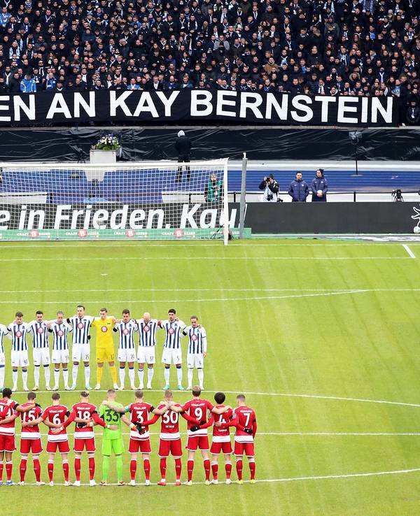 Die Mannschaft der Fortuna Düsseldorf steht gemeinsam mit der Mannschaft von Hertha BSC auf dem Platz. Im Hintergrund die Fans und ein Banner zu Ehren von Kay Bernstein.