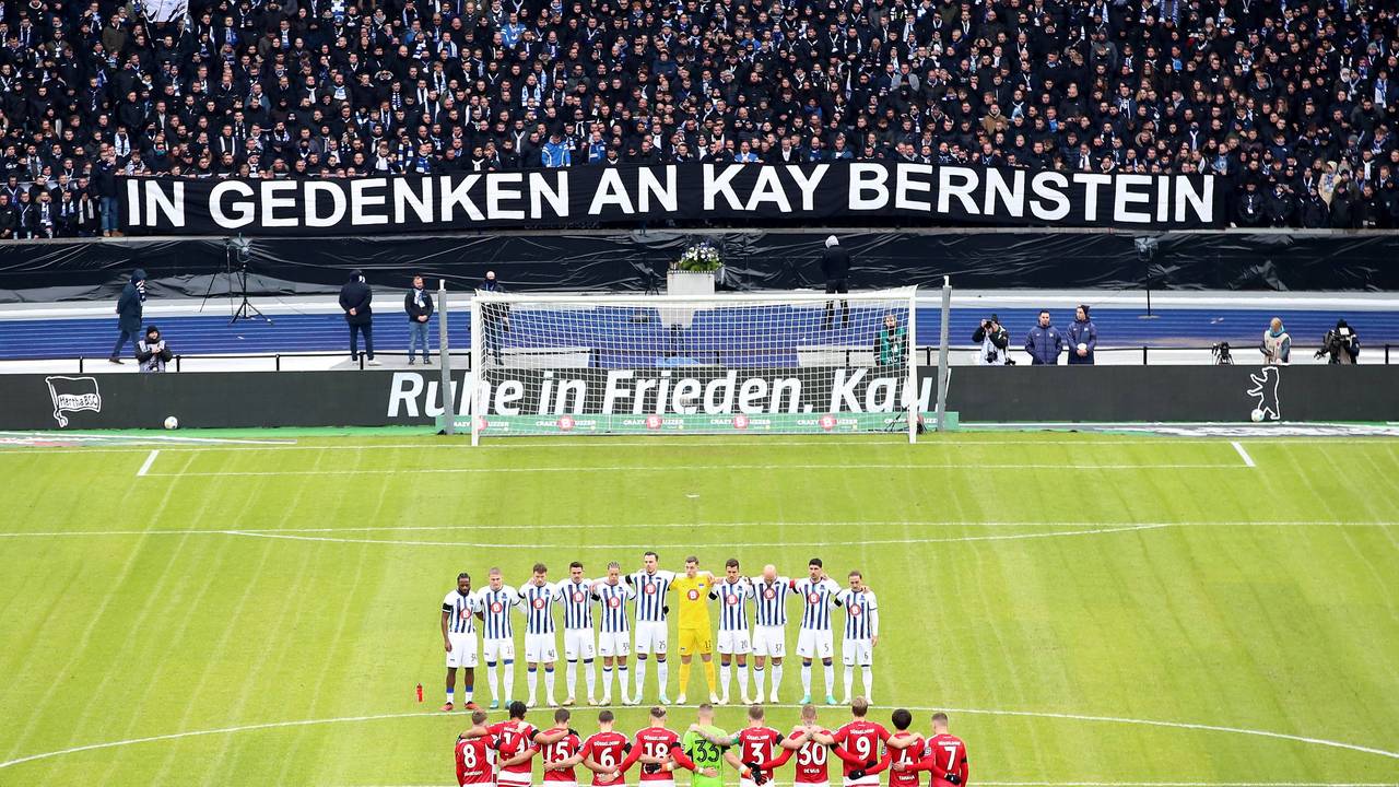 Die Mannschaft der Fortuna Düsseldorf steht gemeinsam mit der Mannschaft von Hertha BSC auf dem Platz. Im Hintergrund die Fans und ein Banner zu Ehren von Kay Bernstein.