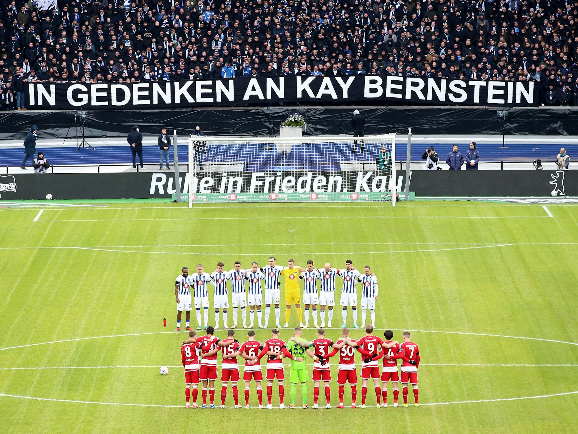 Die Mannschaft der Fortuna Düsseldorf steht gemeinsam mit der Mannschaft von Hertha BSC auf dem Platz. Im Hintergrund die Fans und ein Banner zu Ehren von Kay Bernstein.