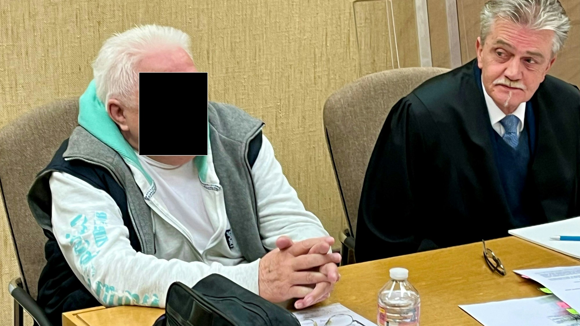 Ein Mann sitzt neben seinem Anwalt auf der Anklagebank, vor sich eine Herrenhandtasche.