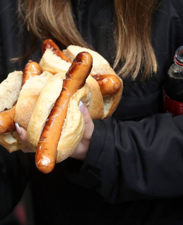 Eine Frau trägt mehrere Bratwürstchen, einen Becher Bier und eine Flasche Cola bei einem Bundesliga-Spiel.