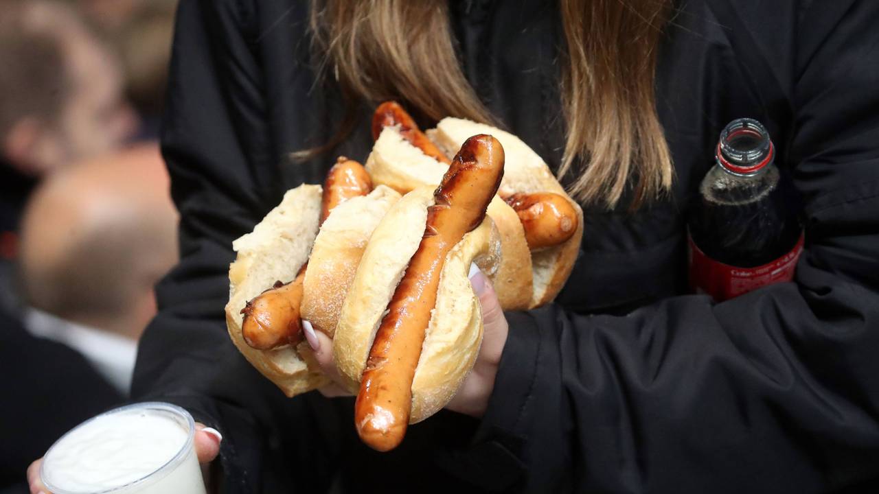 Eine Frau trägt mehrere Bratwürstchen, einen Becher Bier und eine Flasche Cola bei einem Bundesliga-Spiel.