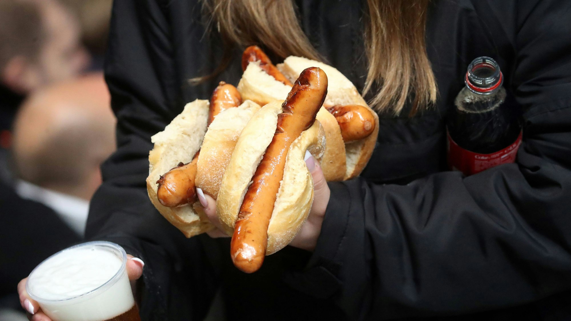 Eine Frau trägt mehrere Bratwürstchen, einen Becher Bier und eine Flasche Cola bei einem Bundesliga-Spiel.