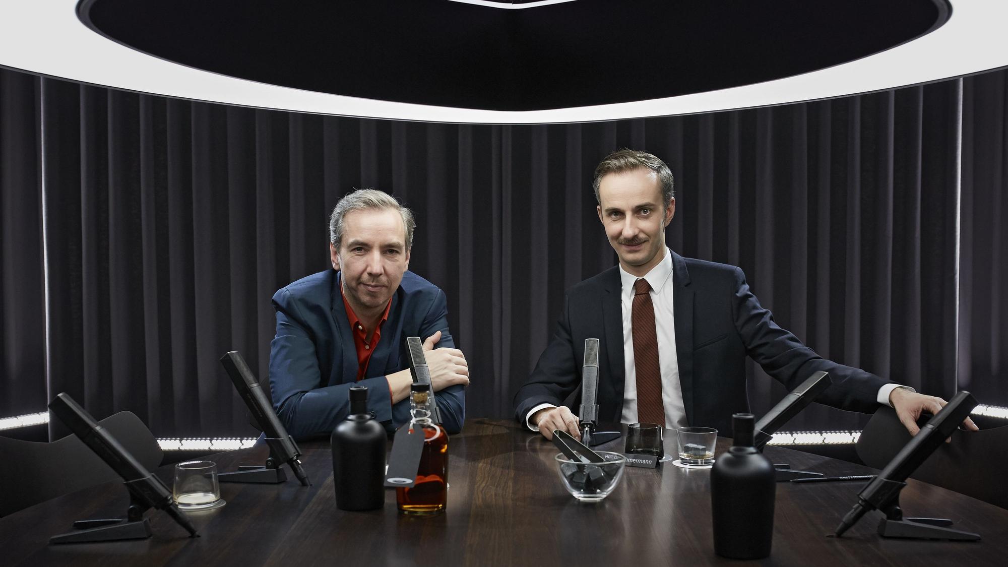 Jan Böhmermann und Olli Schulz sitzen an einem Tisch und blicken in die Kamera.