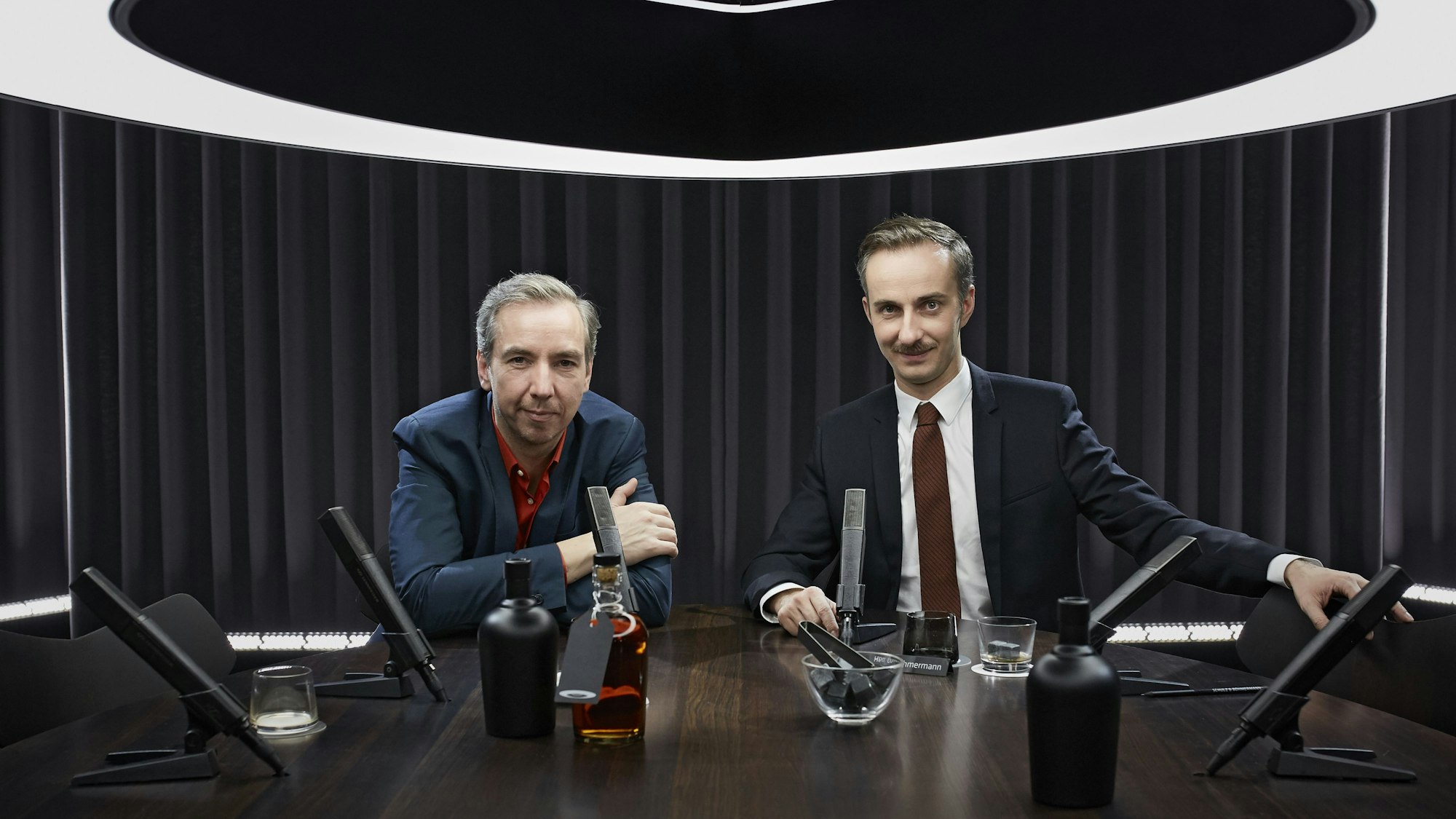 Jan Böhmermann und Olli Schulz sitzen an einem Tisch und blicken in die Kamera.