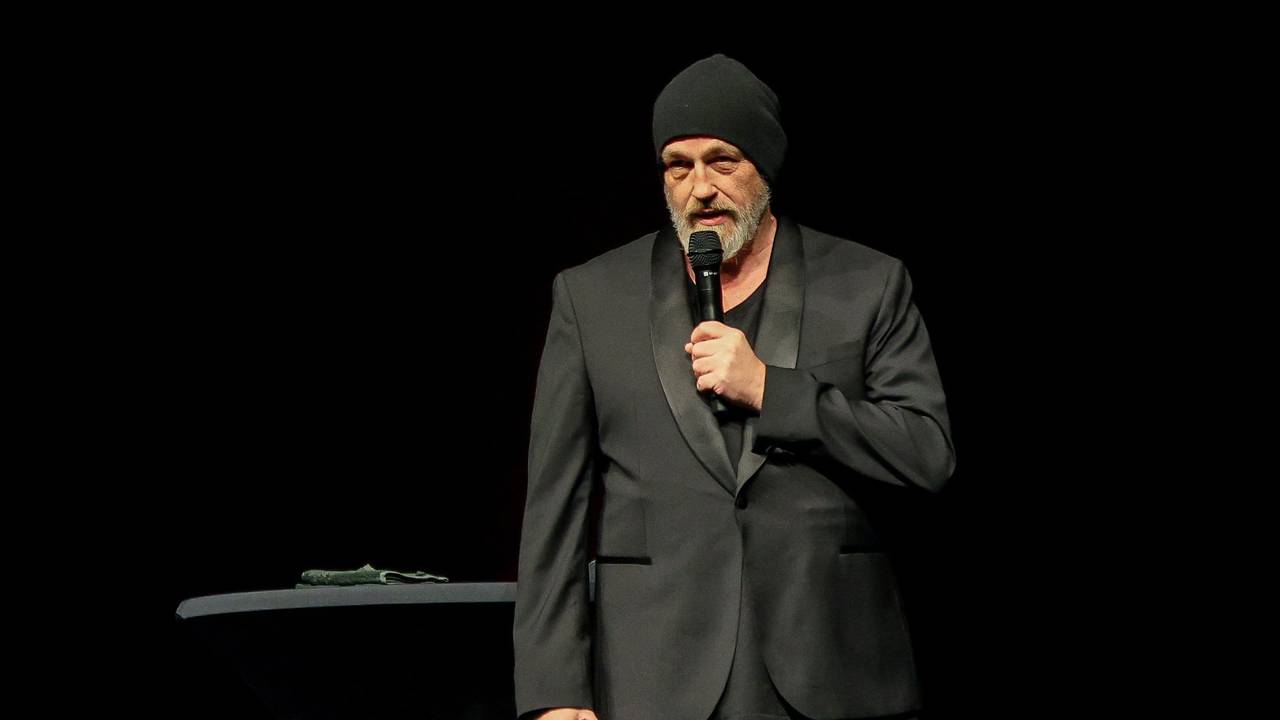 Ein Mann ganz in Schwarz gekleidet und mit einer schwarzen Mütze hält sich ein Mikrofon an den Mund.