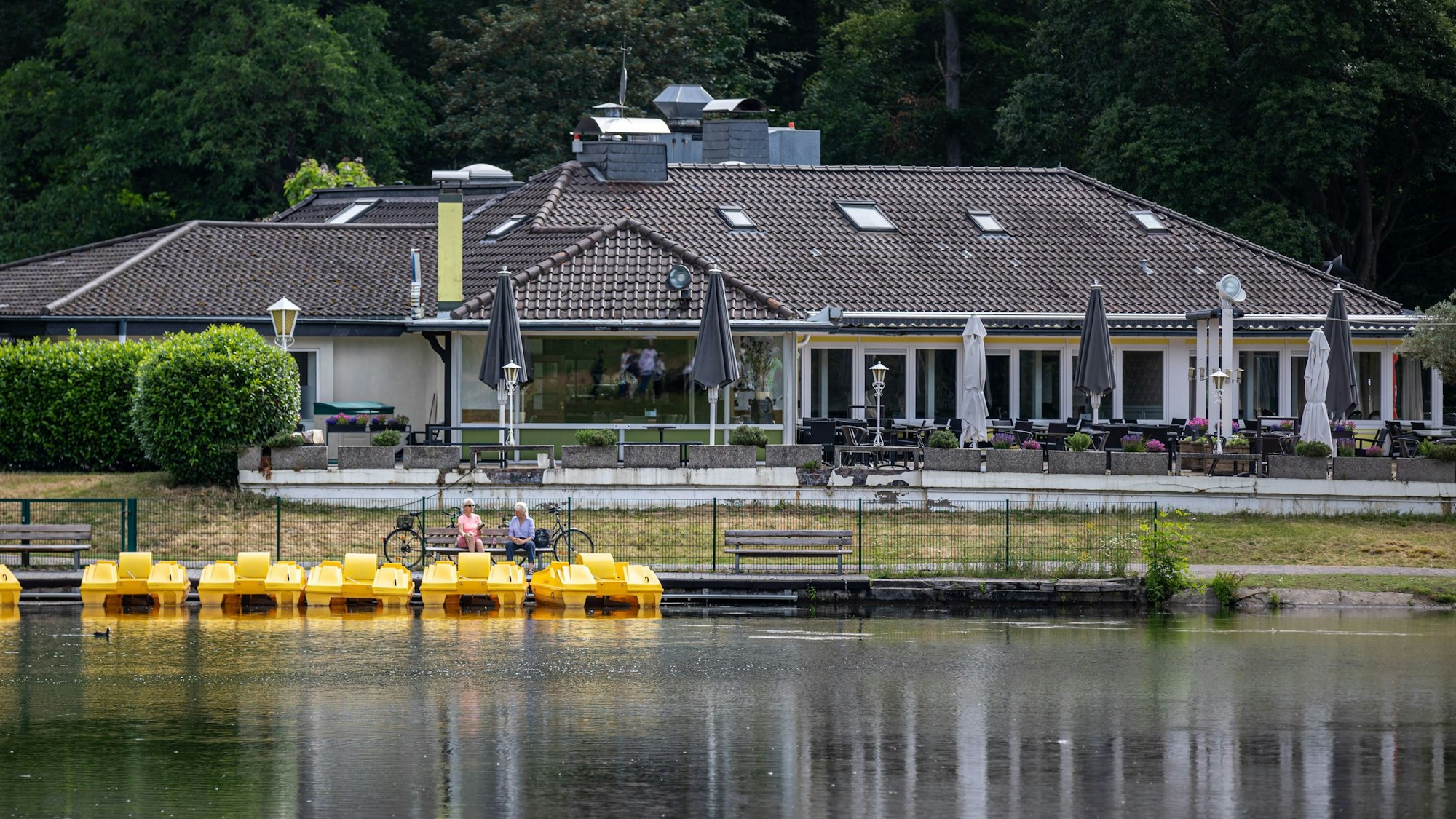Zwei Personen sitzen auf einer Bank am Tretbootverleih am Decksteiner Weiher während im Hintergrund das Restaurant „Haus am See“ zu sehen ist.