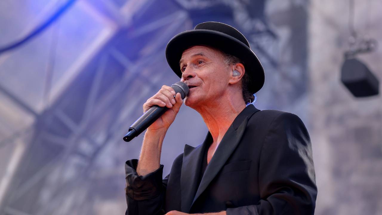 Peter Brings steht mit schwarzem Anzug und schwarzem Hut auf der Bühne und singt in ein Mikrofon.