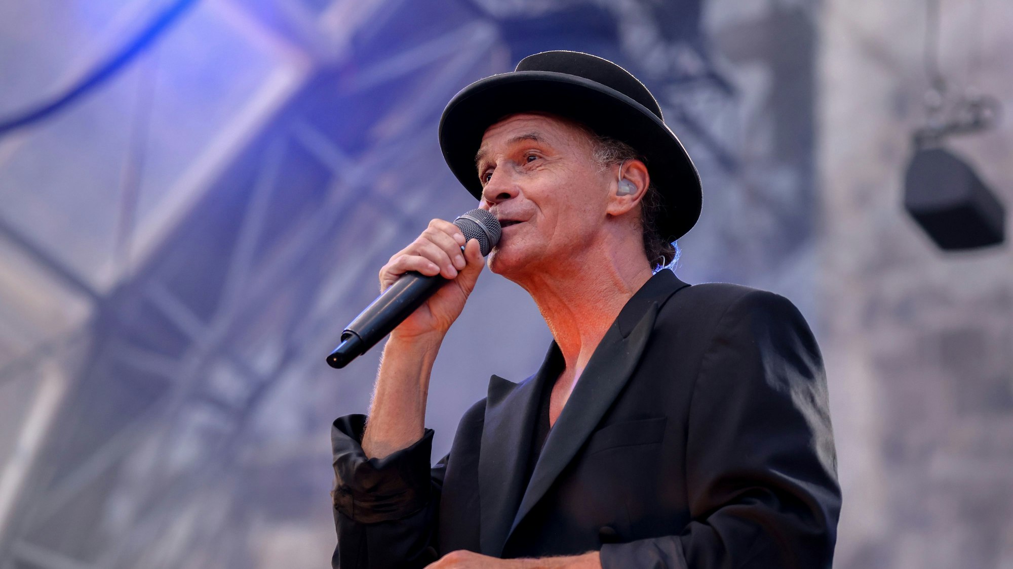 Peter Brings steht mit schwarzem Anzug und schwarzem Hut auf der Bühne und singt in ein Mikrofon.