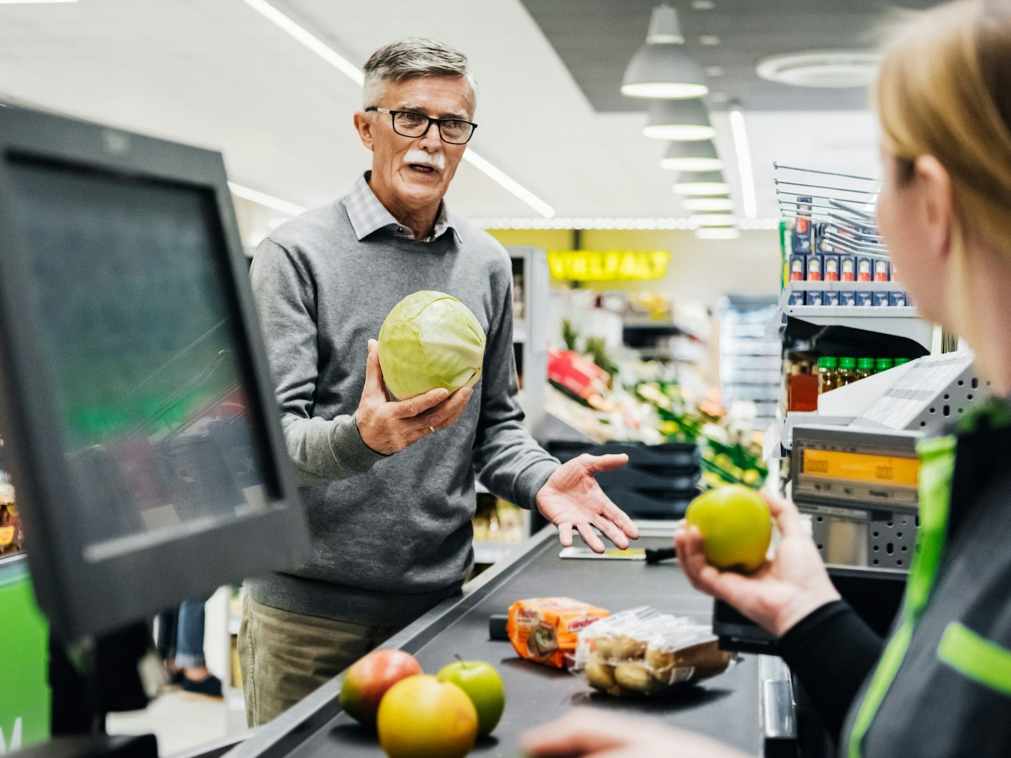 Ein älterer Mann diskutiert an der Kasse mit einer Kassiererin.