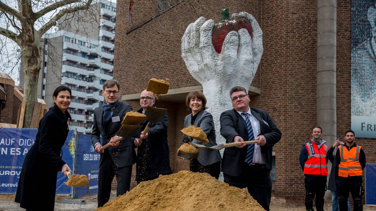 Eine Gruppe von Menschen steht mit Schaufeln vor einem kleinen Hügel Sand.