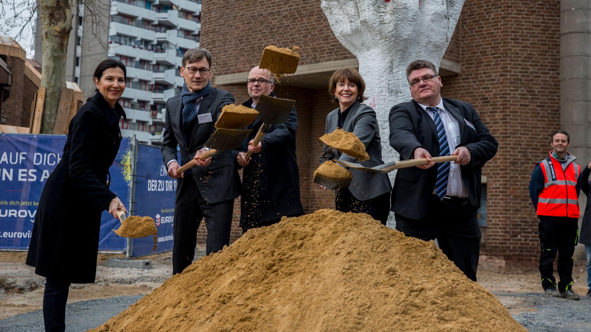 Eine Gruppe von Menschen steht mit Schaufeln vor einem kleinen Hügel Sand.