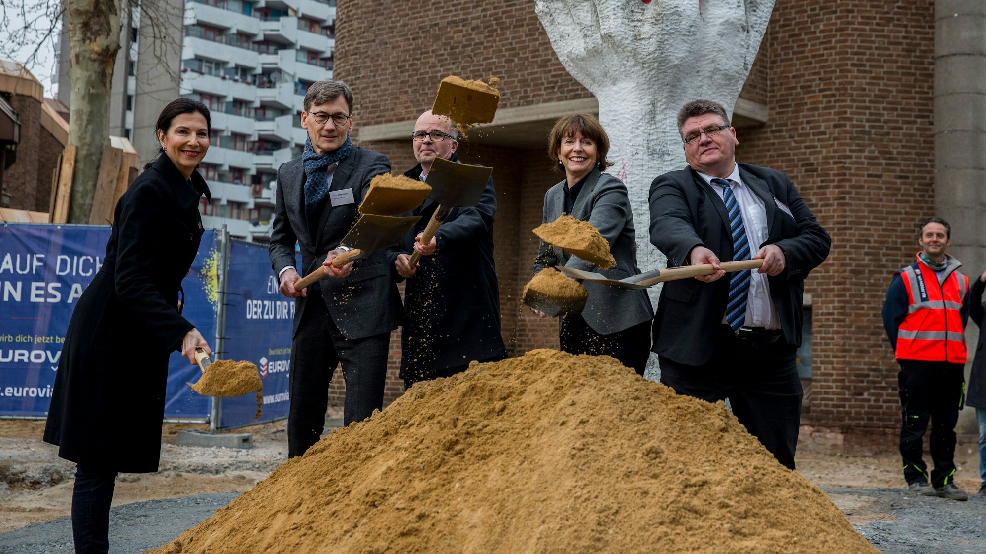 Eine Gruppe von Menschen steht mit Schaufeln vor einem kleinen Hügel Sand.