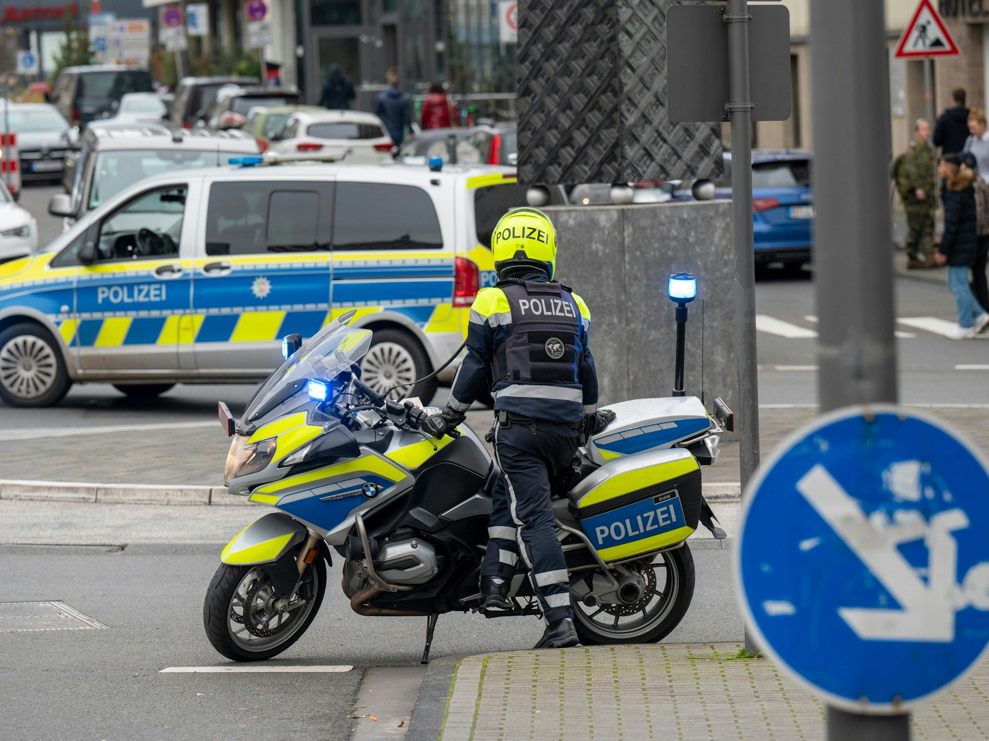 Ein Polizist mit einem Motorrad hat an einem Platz angehalten, ein paar Meter weiter steht ein Polizeiwagen.