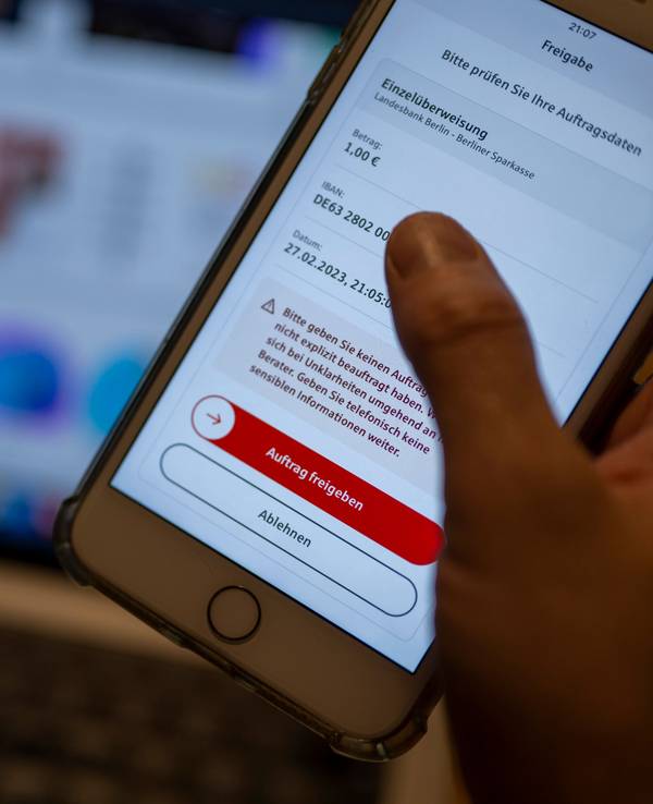 Ein Mann überweist in einer Banking-App auf einem Smartphone Geld, während im Hintergrund auf einem Laptop die Seite eines Online-Handels angezeigt wird, hier im Februar 2023 in Berlin.