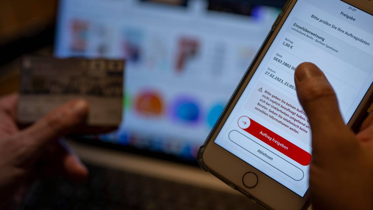 Ein Mann überweist in einer Banking-App auf einem Smartphone Geld, während im Hintergrund auf einem Laptop die Seite eines Online-Handels angezeigt wird, hier im Februar 2023 in Berlin.