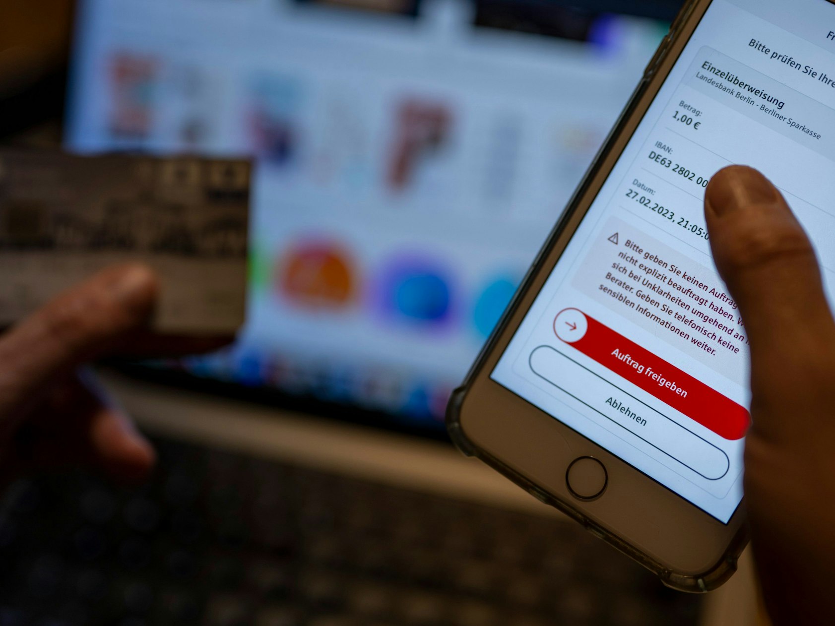 Ein Mann überweist in einer Banking-App auf einem Smartphone Geld, während im Hintergrund auf einem Laptop die Seite eines Online-Handels angezeigt wird, hier im Februar 2023 in Berlin.