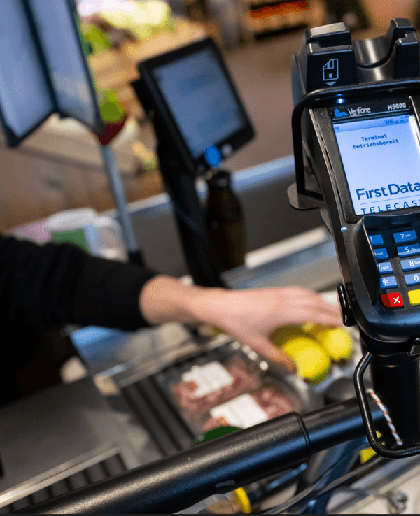 Ein Kassierer scannt in einem Supermarkt an der Kasse die Produkte. Im Vordergrund ist ein EC Terminal zu sehen.