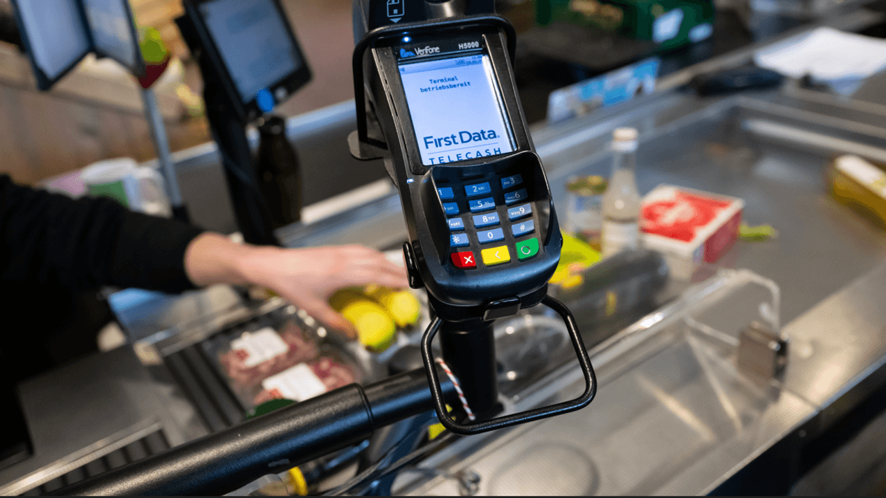 Ein Kassierer scannt in einem Supermarkt an der Kasse die Produkte. Im Vordergrund ist ein EC Terminal zu sehen.