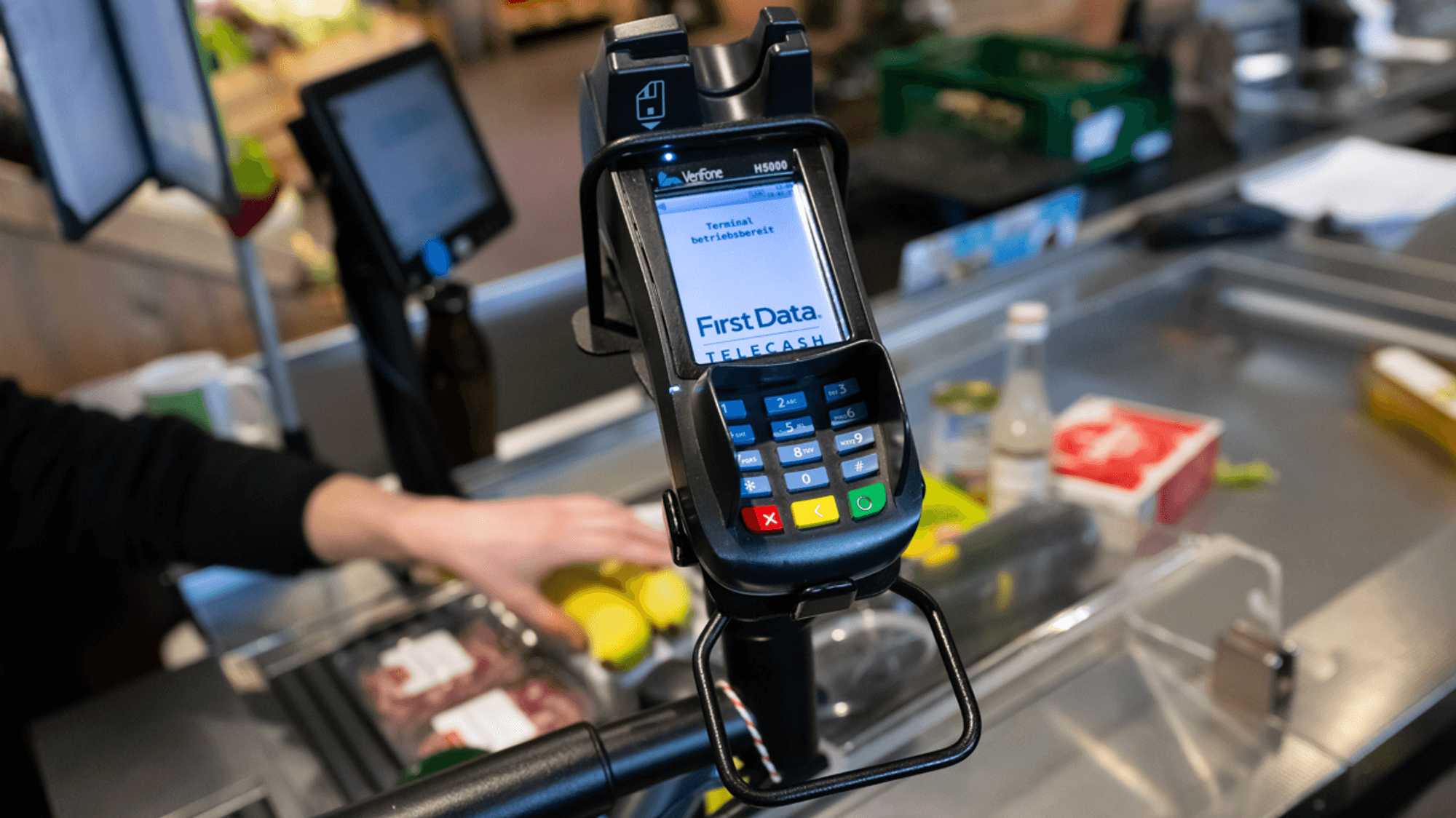Ein Kassierer scannt in einem Supermarkt an der Kasse die Produkte. Im Vordergrund ist ein EC Terminal zu sehen.