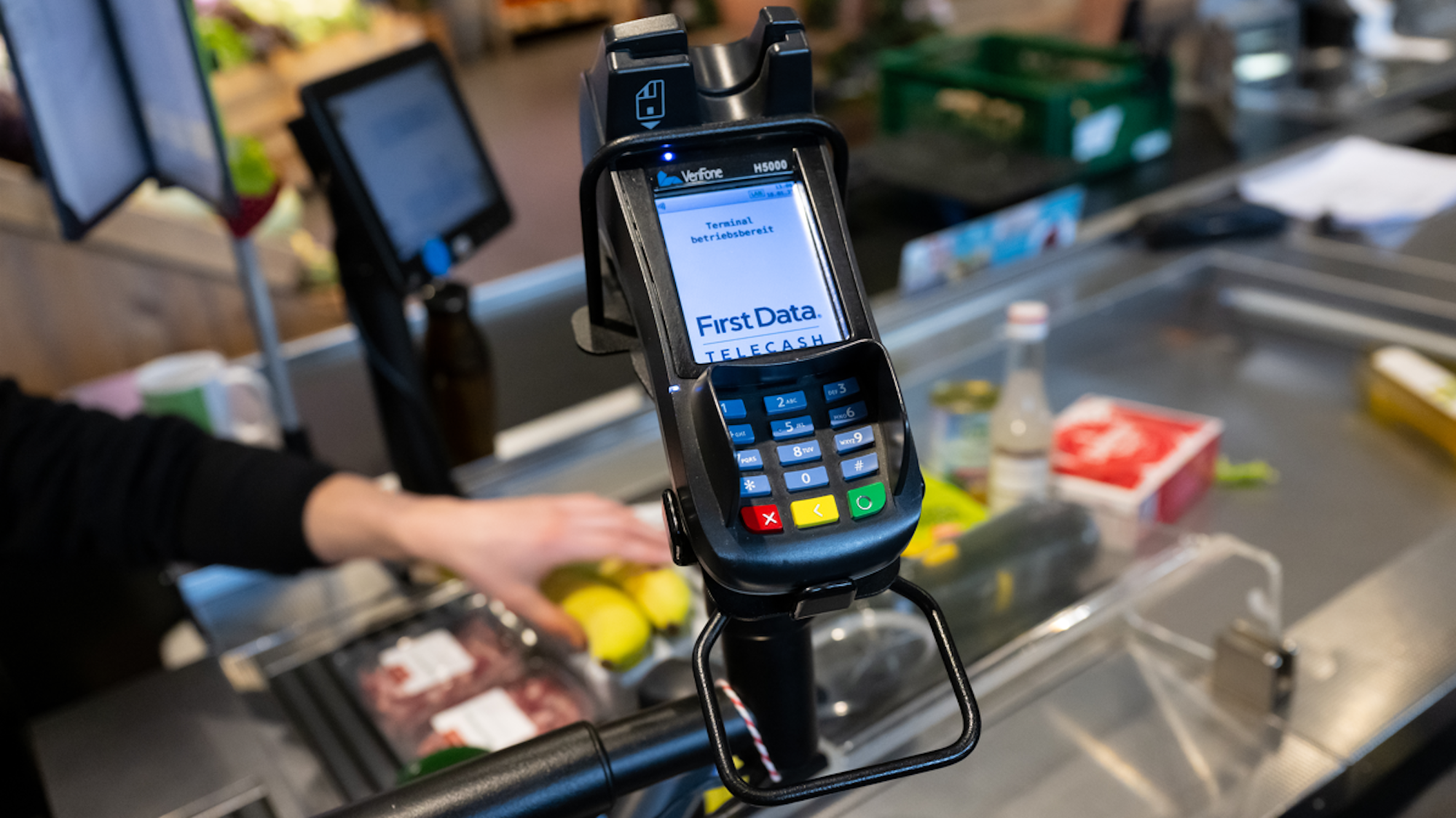 Ein Kassierer scannt in einem Supermarkt an der Kasse die Produkte. Im Vordergrund ist ein EC Terminal zu sehen.