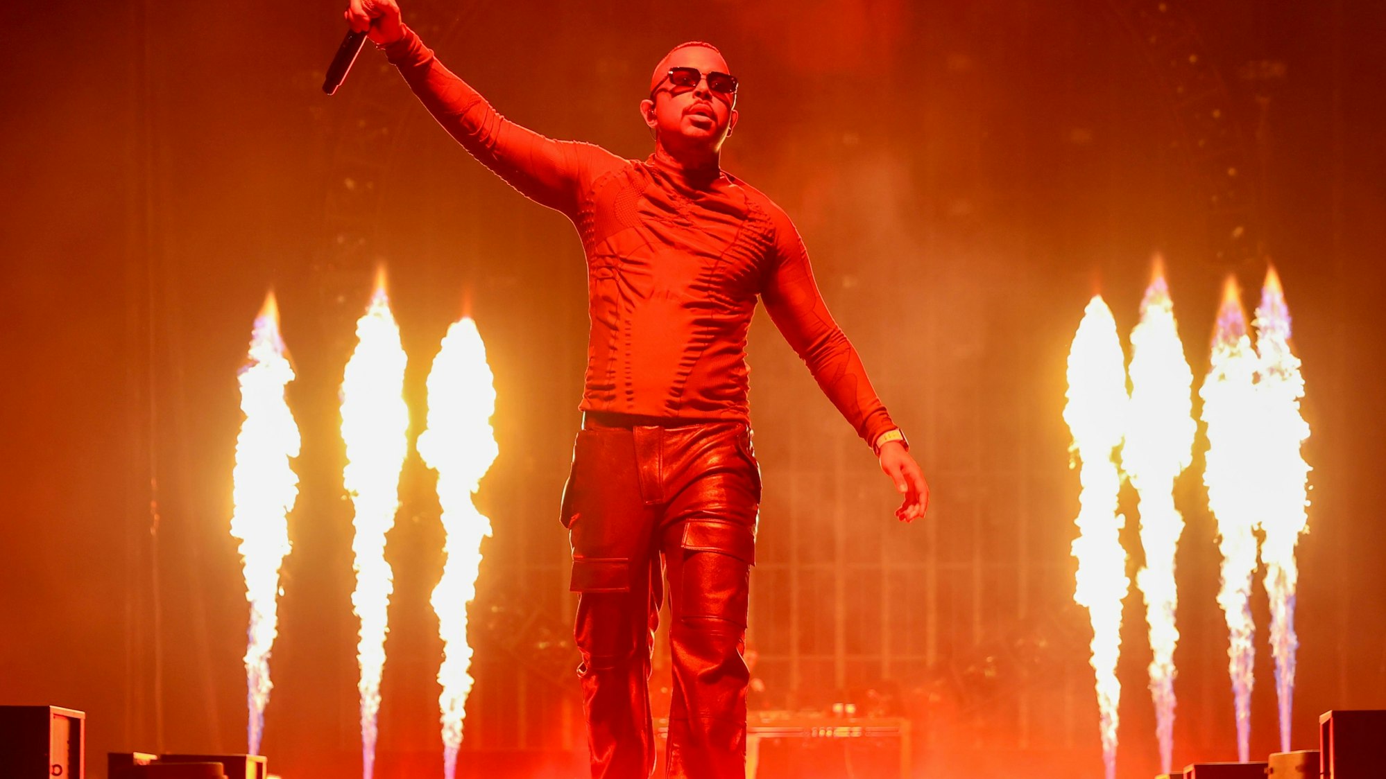 Ein Mann reckt ein Mikrofon in die Höhe, im Hintergrund sind mehrere Flammen.