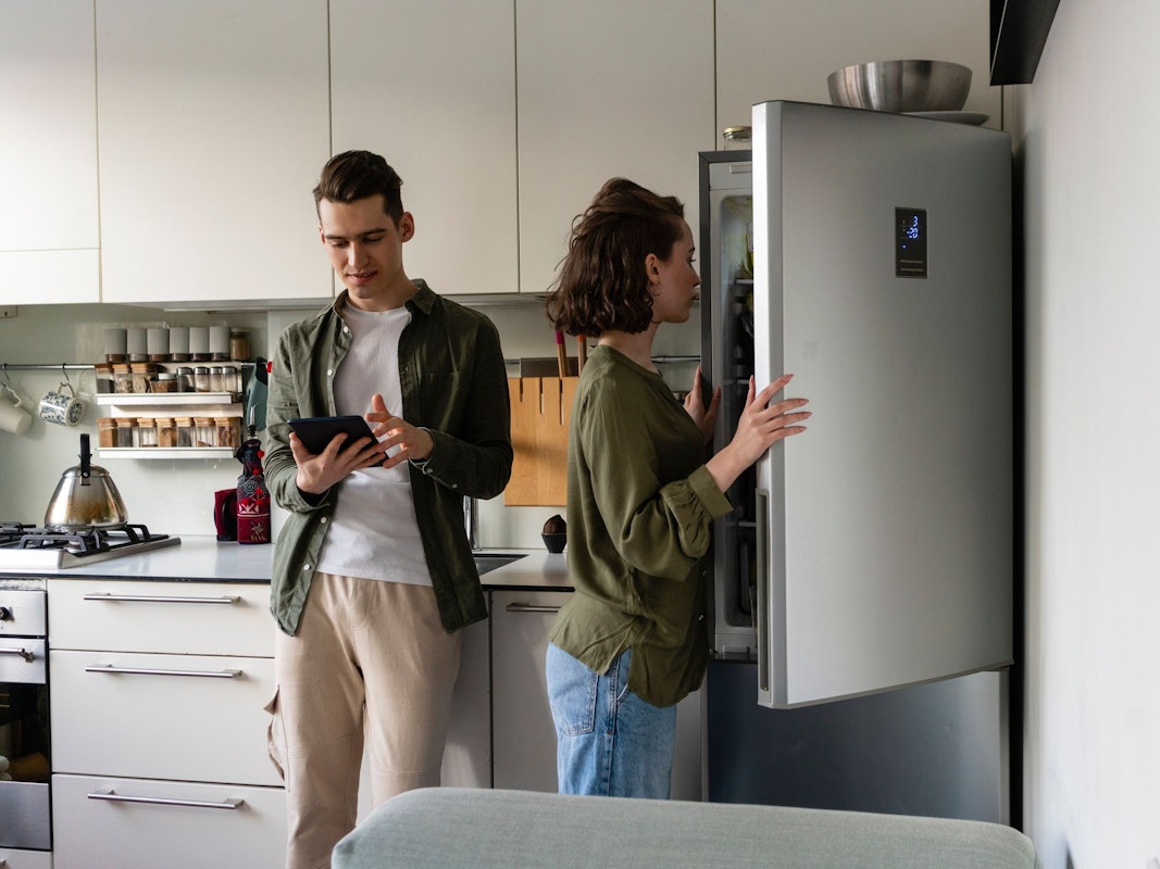 Auf dem Foto sieht man eine Frau einen Kühlschrank öffnen, neben ihr steht ein Mann, welcher auf sein Tablet schaut.